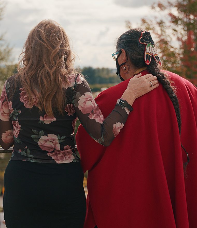 Two women facing away from camera, one of the women is dressed in traditional Native American clothing